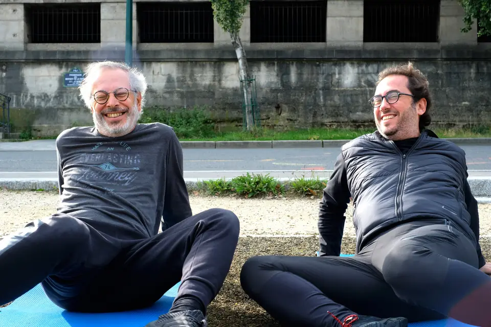 Senior et son partenaire d'entraînement assis sur des tapis, riant pendant une séance au bord de la Seine